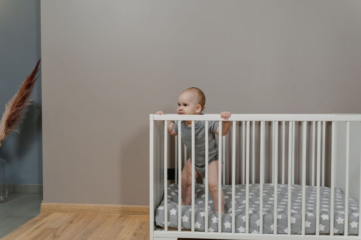Enfant dans une berceuse blanche, mains sur les barreaux, sol en bois, mur gris
